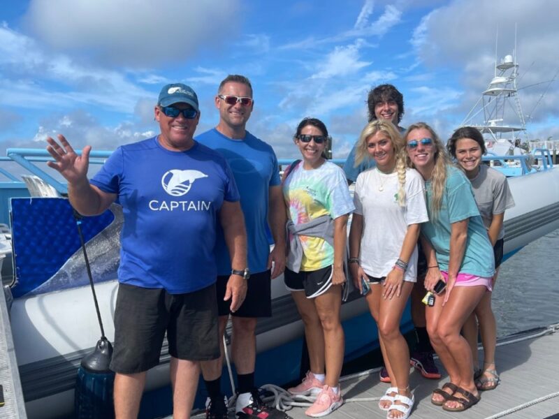 A group of seven people stand smiling on a dock in front of a white boat under a blue sky with scattered clouds. One man wears a blue Captain shirt and waves at the camera. Everyone is dressed casually for warm weather. — Blue Wave Adventures