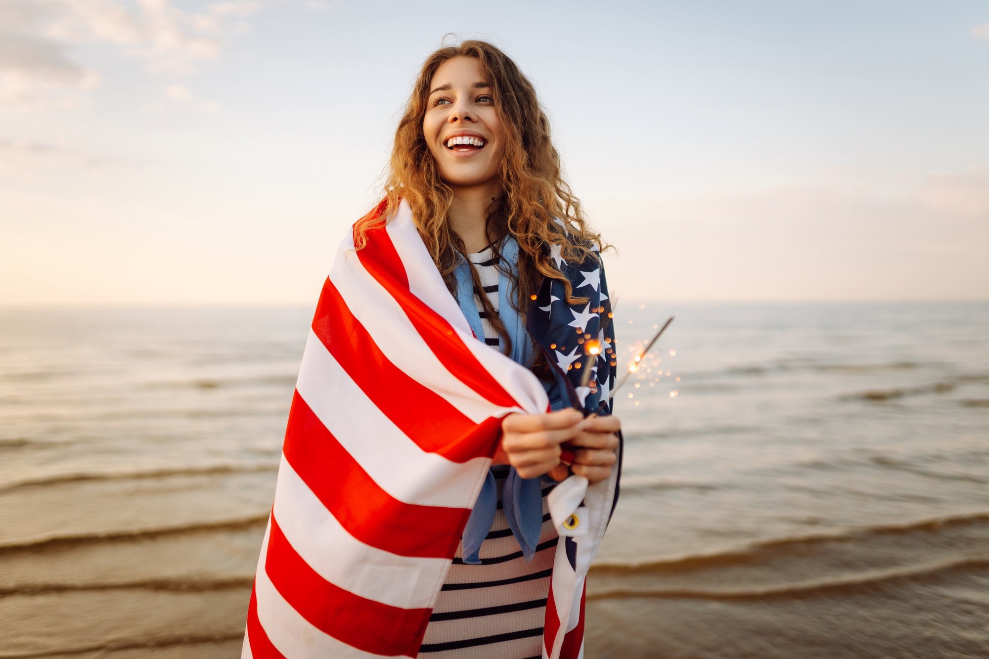 Young woman with American flag and sparklers on beach. Patriotic holiday. USA celebrate 4th of July.